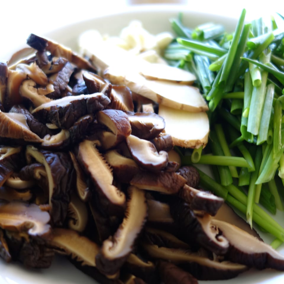 Sliced shiitake mushrooms, ginger, and green onions on a white plate, ready for Chinese cooking.